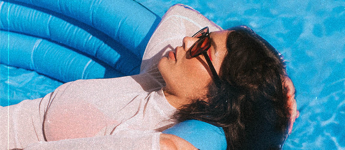 JJ Wilde is shown in profile from the waist up lying in a blue float, in a pool. She's wearing a white long sleeve shirt, sunglasses, and her hair is over the edge of the float, floating in the pool