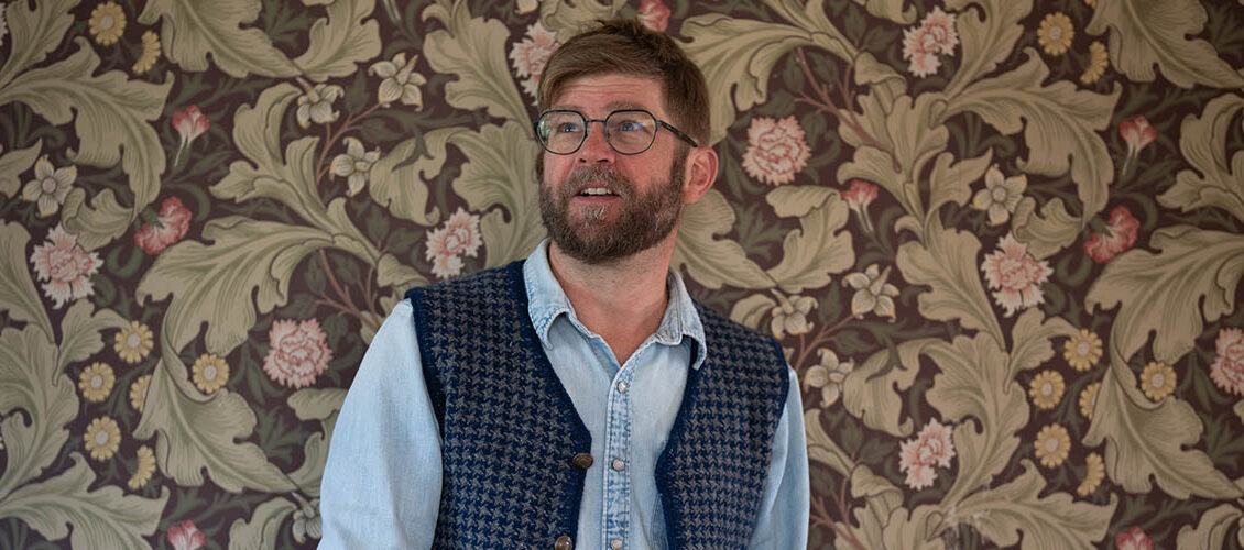 Chris Ludecke is seen from the chest up standing in front of floral wall paper, looking off to the left. He is wearing a chambray shirt and a knit button up vest that is open.
