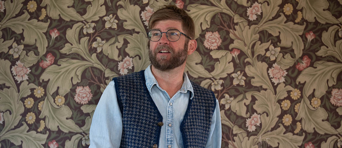 Chris Ludecke is seen from the chest up standing in front of floral wall paper, looking off to the left. He is wearing a chambray shirt and a knit button up vest that is open.